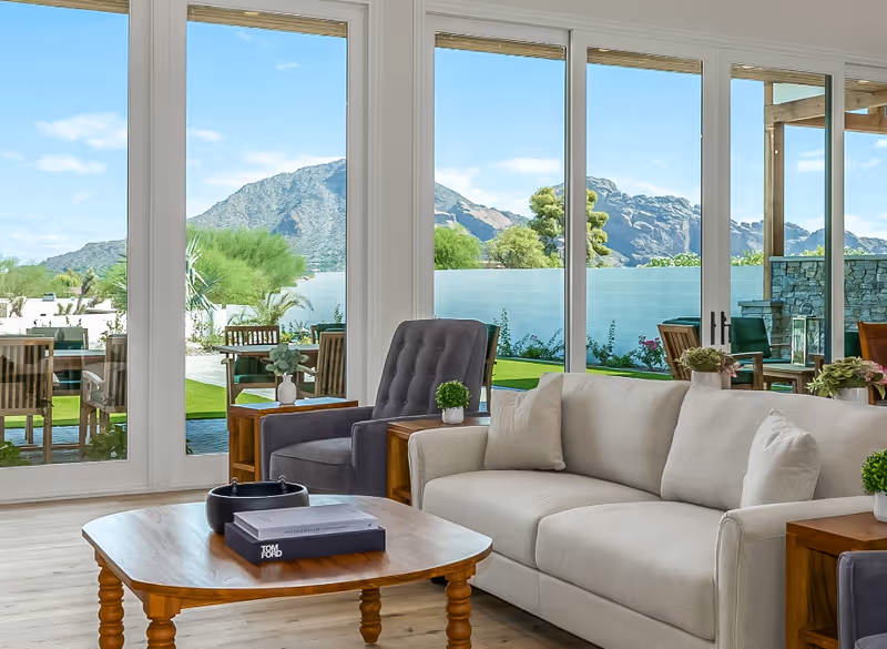 A bright living room with a beige sofa, a gray armchair, wooden side tables with small potted plants, and a wooden coffee table with books and a decorative bowl. Large glass doors provide a view of an outdoor patio with wooden chairs and tables, green lawn, and mountains in the background under a blue sky.