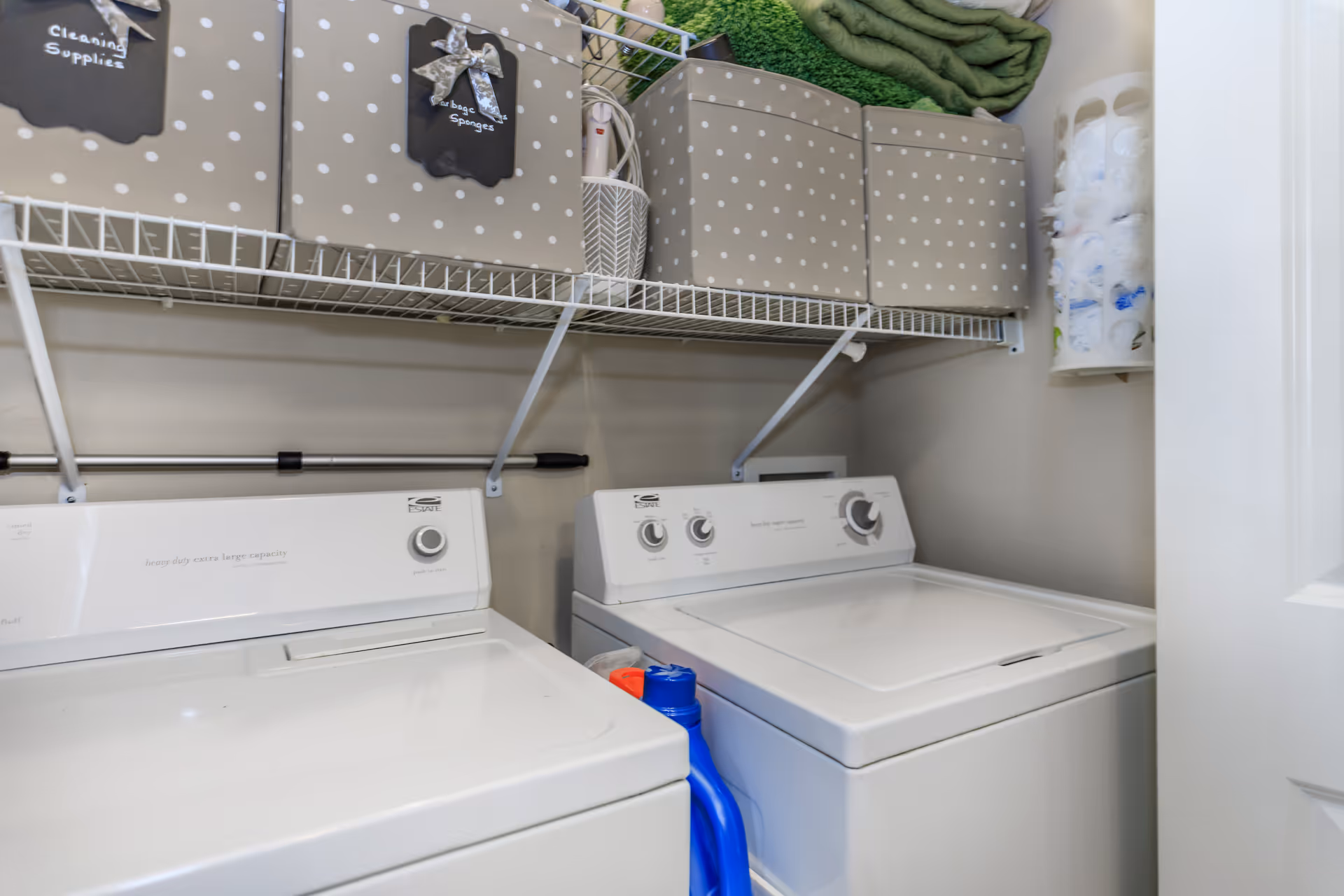 A small laundry room with side-by-side washer and dryer, wire shelving holding polka-dot storage boxes and laundry supplies.
