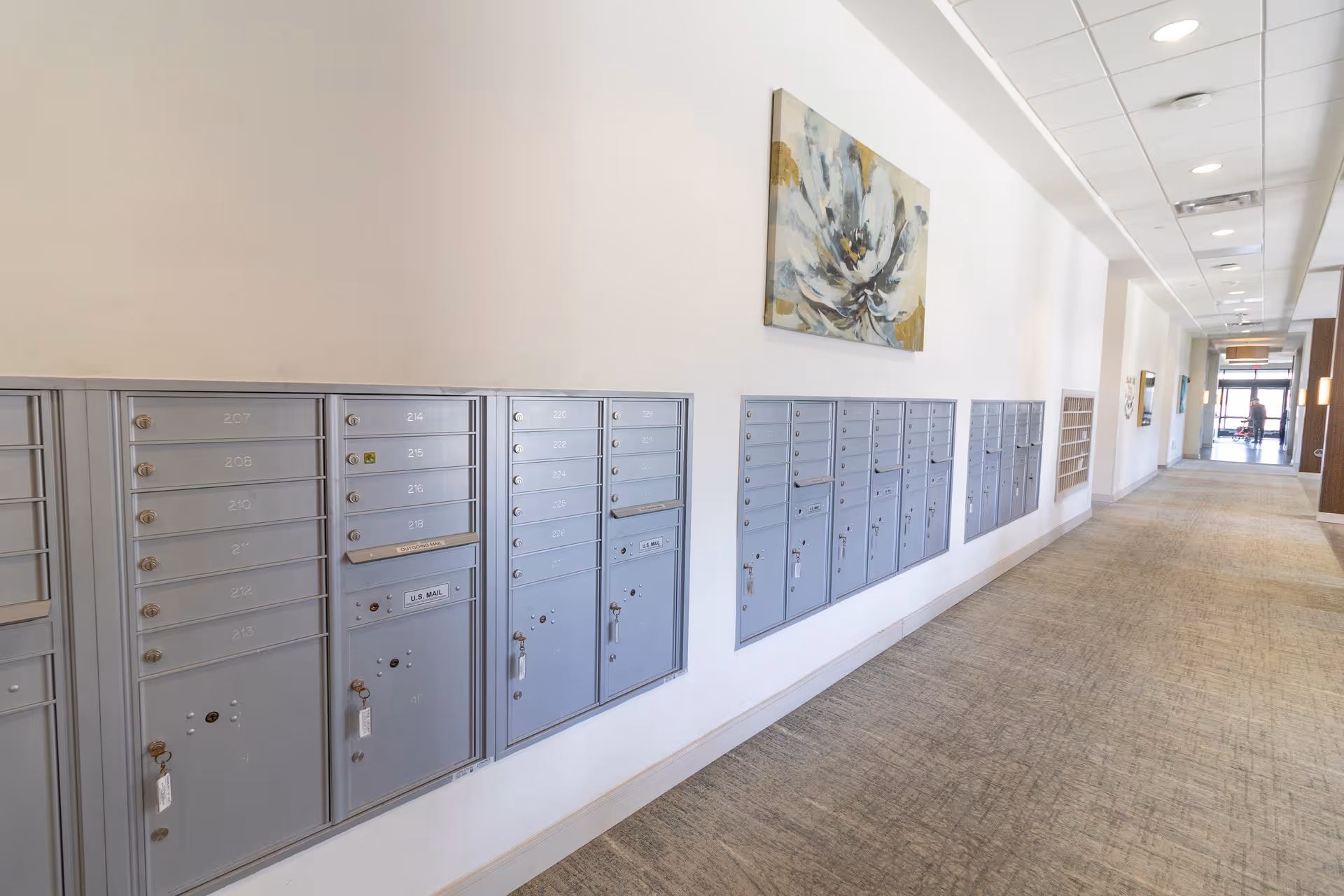 A long hallway with multiple rows of gray mailboxes mounted on the left wall. Above the mailboxes is a large floral painting. The hallway has a light-colored carpet and a white ceiling with recessed lighting. At the far end of the hallway, a person is visible near the entrance or exit door.