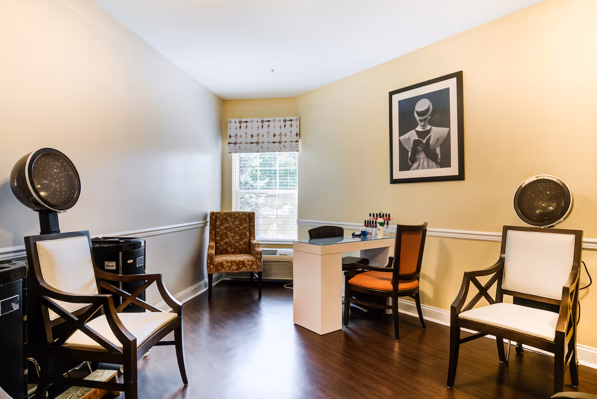 A small salon room with two vintage hair dryers positioned over two wooden chairs with white cushions, a glass-top manicure table with two chairs, and a window with a patterned valance letting in natural light. A framed black and white photograph hangs on the beige wall.