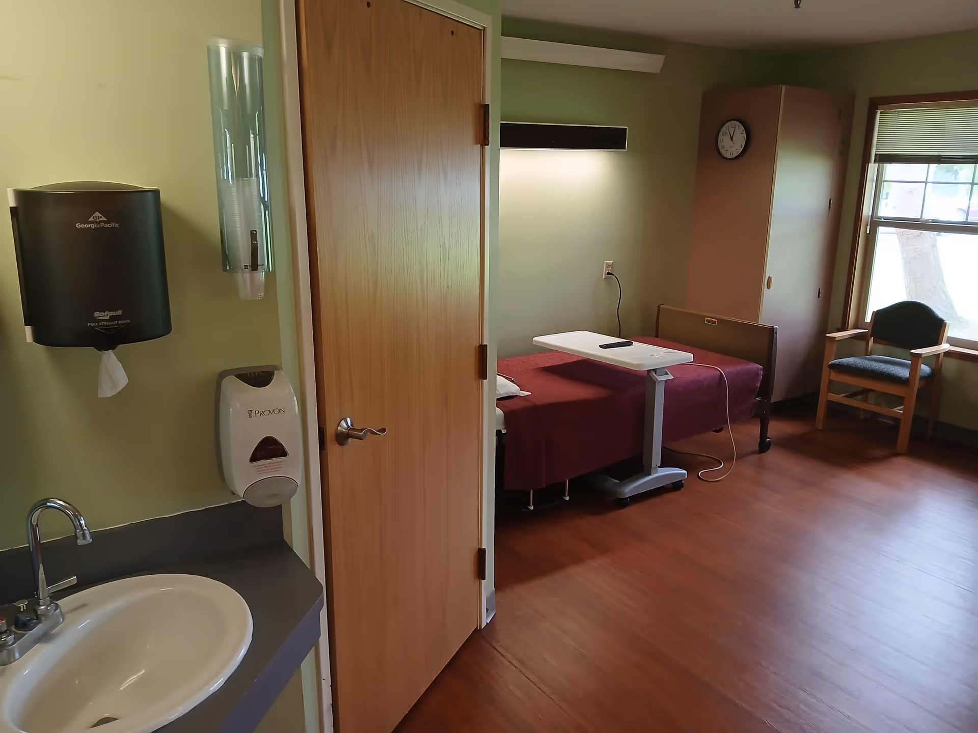 A room with a single bed covered with a maroon blanket, a small adjustable overbed table with a remote on it, a wooden chair near a window, a wall clock, a wooden cabinet, and a sink with soap and paper towel dispensers mounted on the wall.