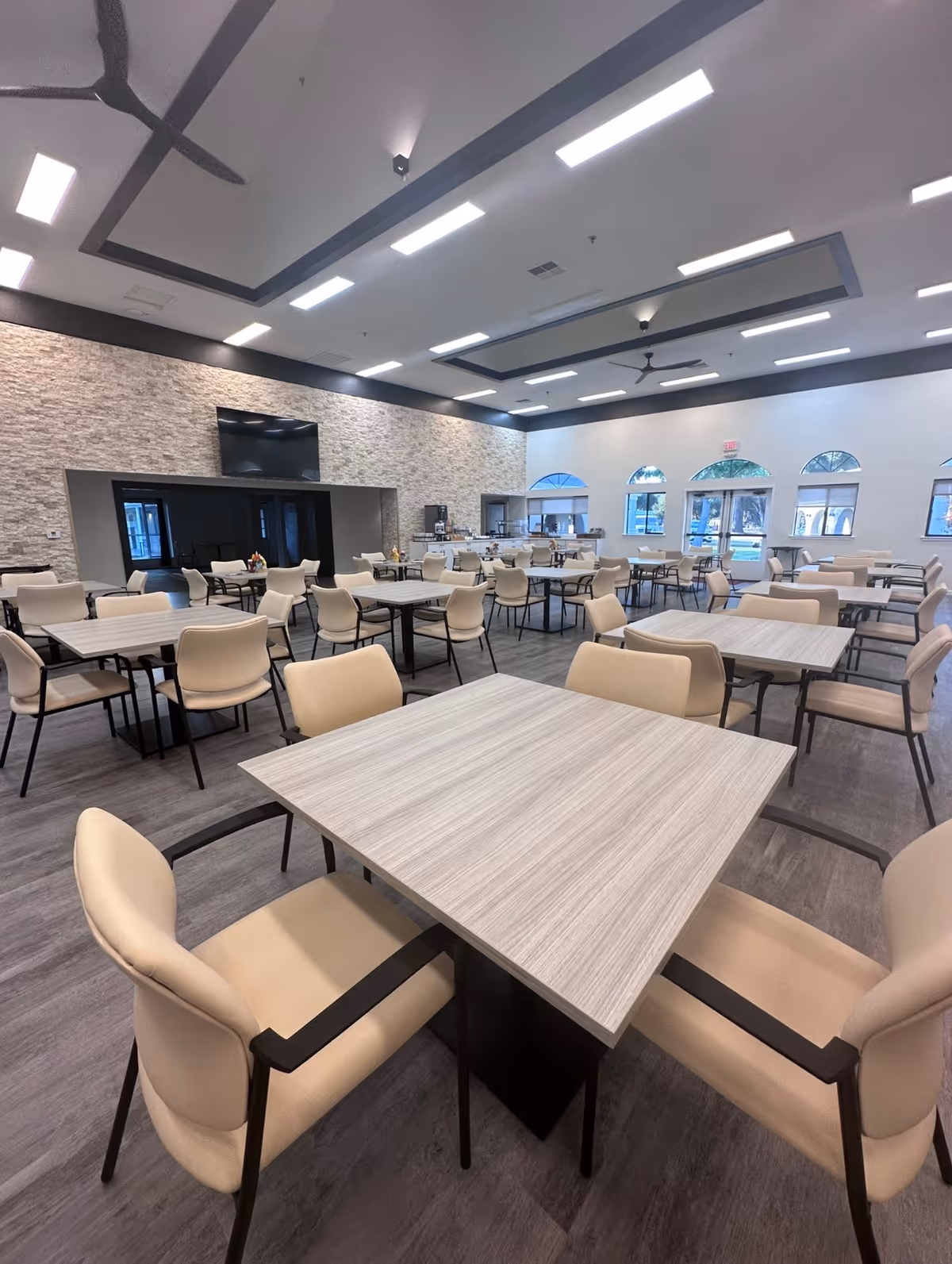 A spacious dining room in Oakview Village Independent Senior Living with multiple square tables and beige cushioned chairs arranged neatly. The room features a modern design with a stone accent wall, a large flat-screen TV mounted on the wall, and several arched windows letting in natural light. The ceiling has recessed lighting and ceiling fans, and the floor is covered with wood-style flooring.