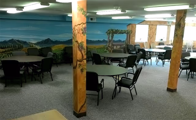 Interior view of a common area with multiple round tables and chairs arranged throughout the room. The walls feature a mural depicting a landscape with mountains and greenery. Several pillars are painted with vine and grape designs. The room is well-lit with ceiling lights and has carpeted flooring.