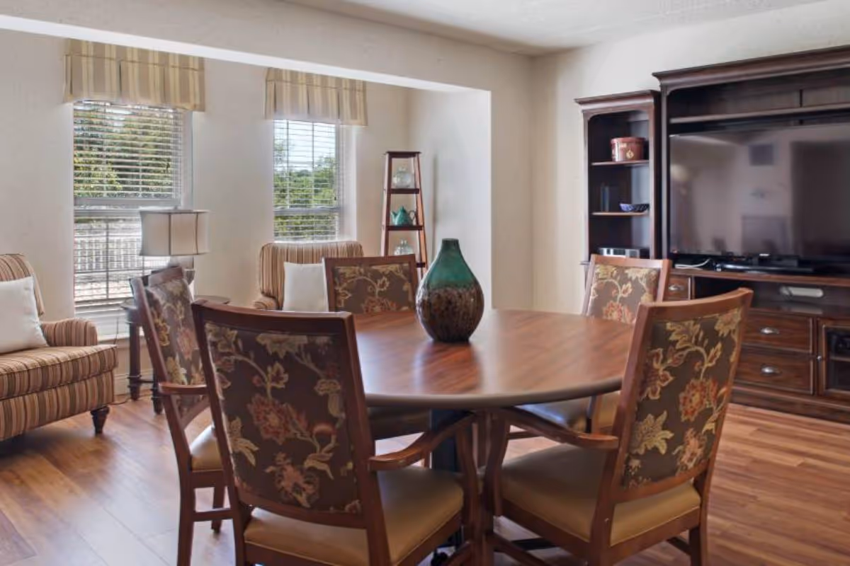 A bright communal room with a round wooden dining table surrounded by upholstered chairs, armchairs by windows, and a TV cabinet.
