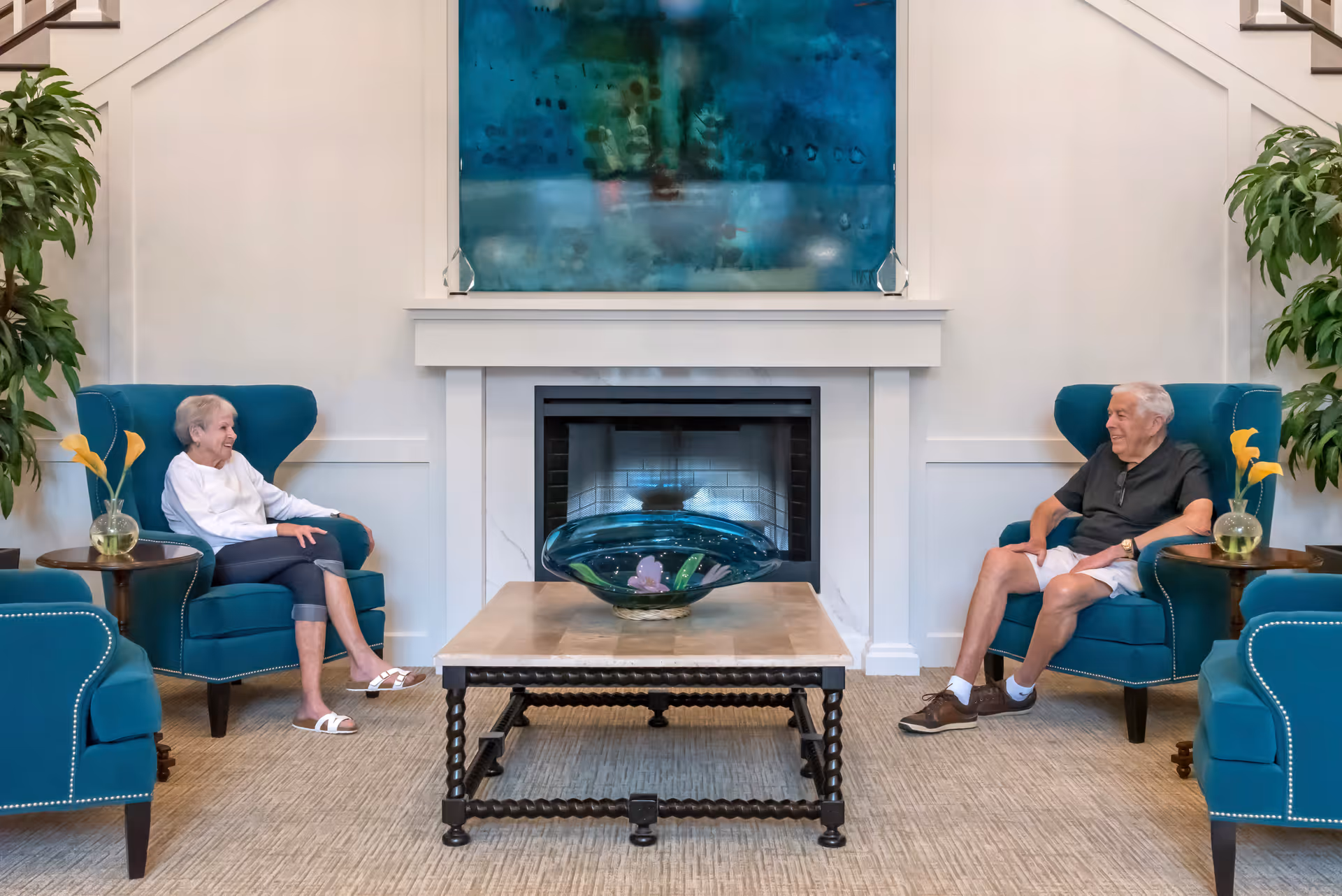 Two older adults sit facing each other in teal armchairs across from a coffee table and fireplace in a lounge.