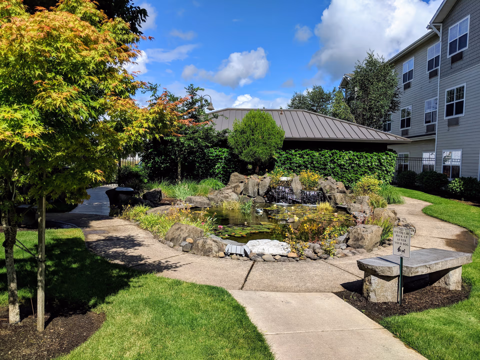 A landscaped outdoor area at Timber Pointe Senior Living featuring a small pond with a rock waterfall, surrounded by greenery and plants. There is a paved walkway curving around the pond, a stone bench with a sign reminding visitors to pick up after their dogs, and a multi-story building in the background under a partly cloudy blue sky.