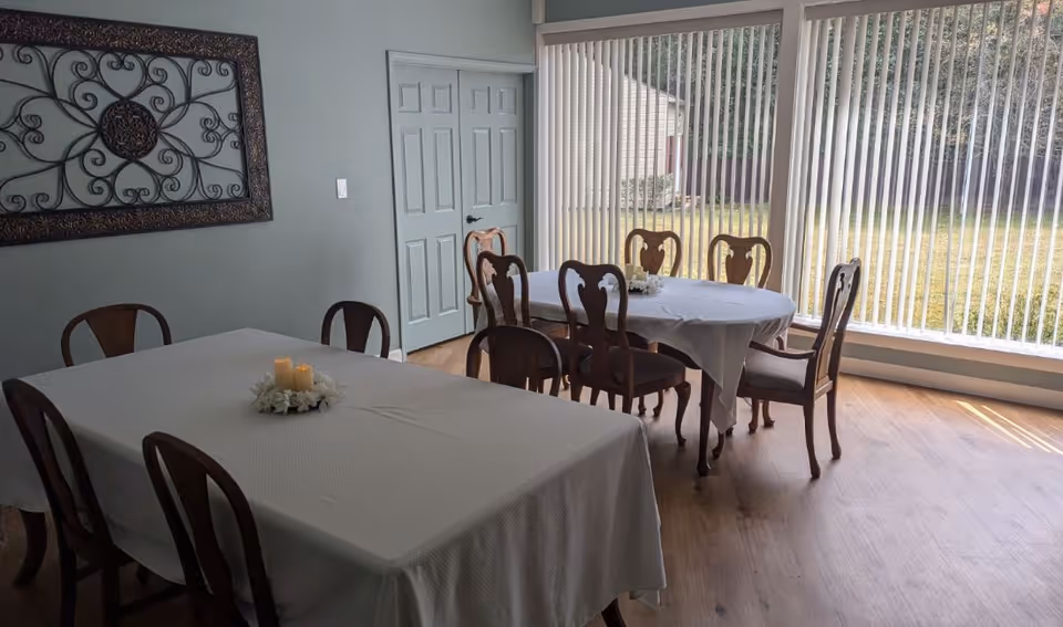 Dining room with two white-clothed tables and wooden chairs next to large vertical blinds overlooking a yard.