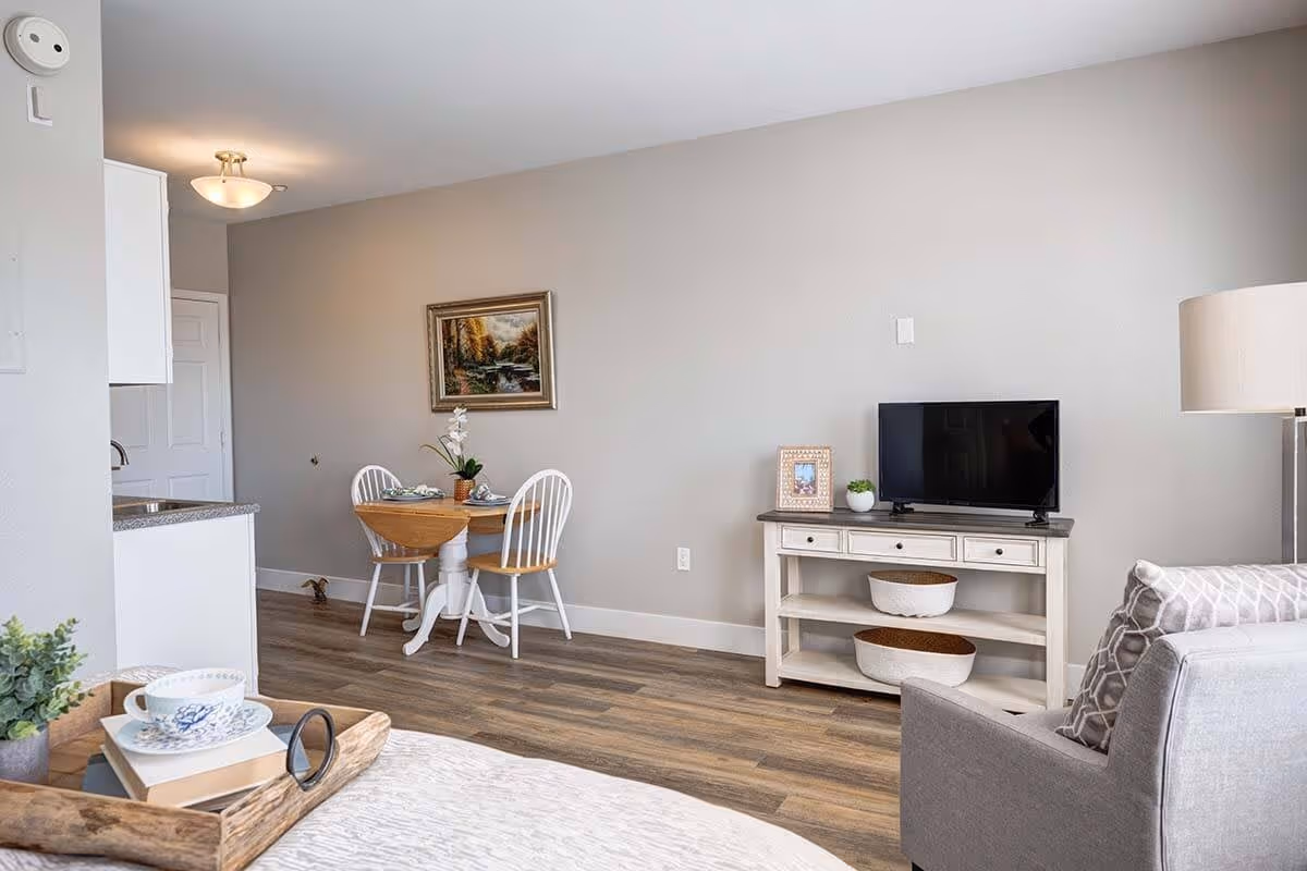 A cozy living area in an assisted living facility featuring a small round dining table with two chairs, a TV on a white console table with baskets underneath, a gray armchair with patterned cushions, and a tray with a cup and books on a bed or sofa. The room has wooden flooring, neutral-colored walls, and a framed landscape painting on the wall.