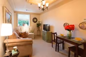 A cozy living room with a beige sofa, a small wooden dining table set for two with red flowers in a vase, a green TV stand with a television, a window with a view of greenery, and warm lighting from a chandelier and a table lamp.