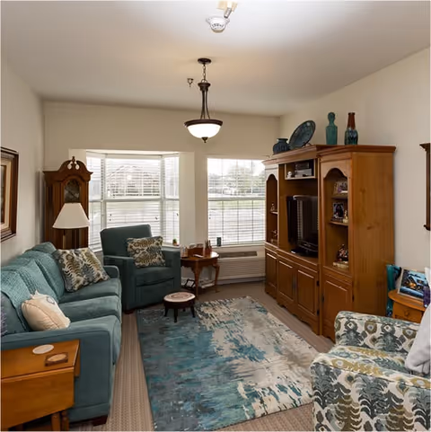 A cozy living room with a teal sofa, matching armchair, patterned armchair, wooden entertainment center with a TV, a grandfather clock, a small wooden side table with a lamp, and a large window with white blinds letting in natural light. The room has a blue and beige area rug on the floor.