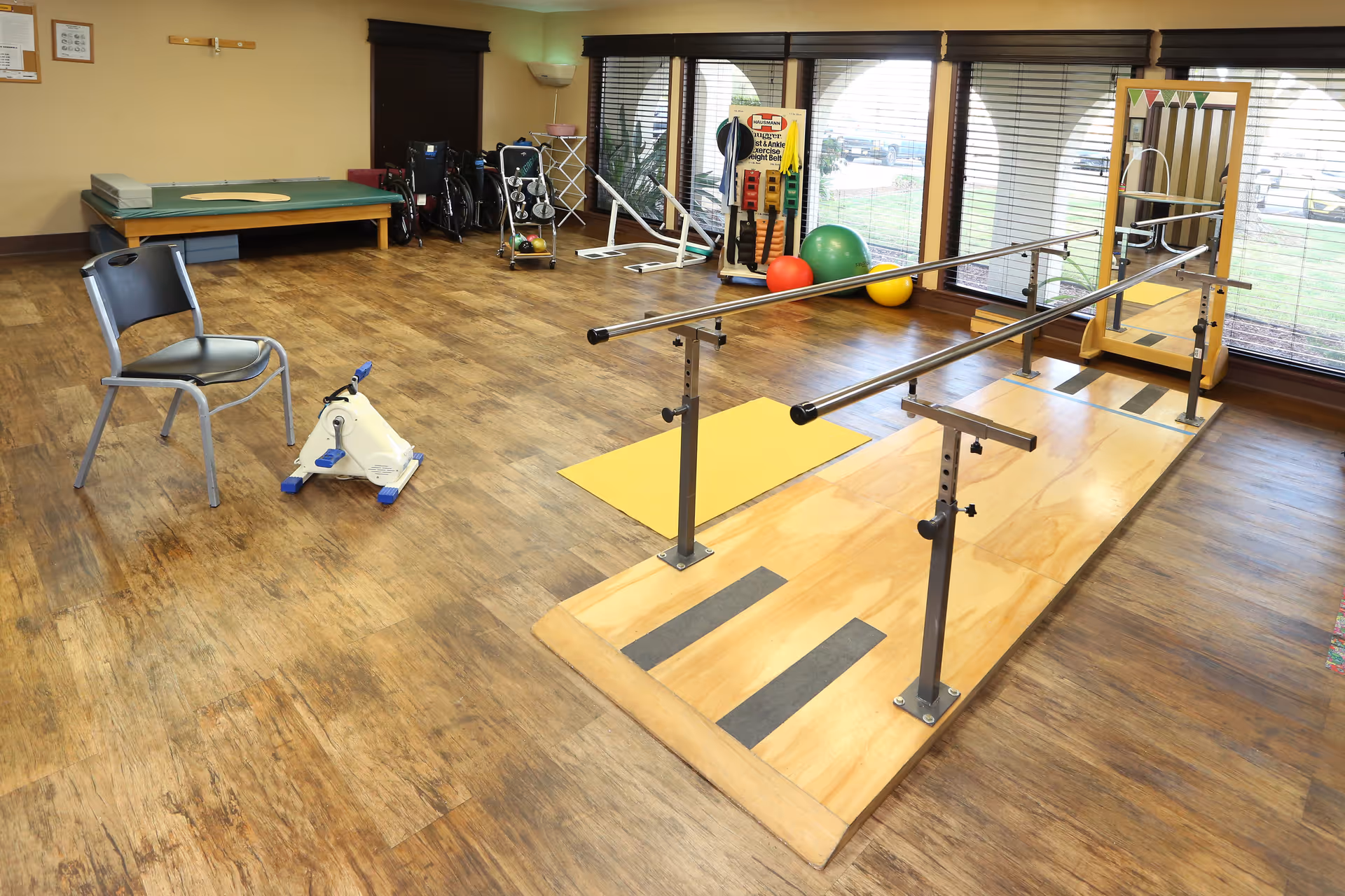 Rehabilitation therapy room with parallel bars, exercise equipment, stability balls, and chairs on wood flooring.