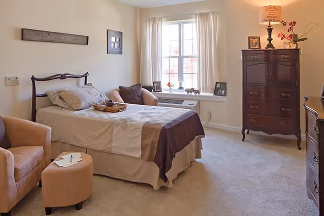 A cozy bedroom with a neatly made bed featuring beige and brown bedding, two armchairs with a small ottoman, a window with white curtains, and a dark wooden dresser with a lamp and decorative items on top.