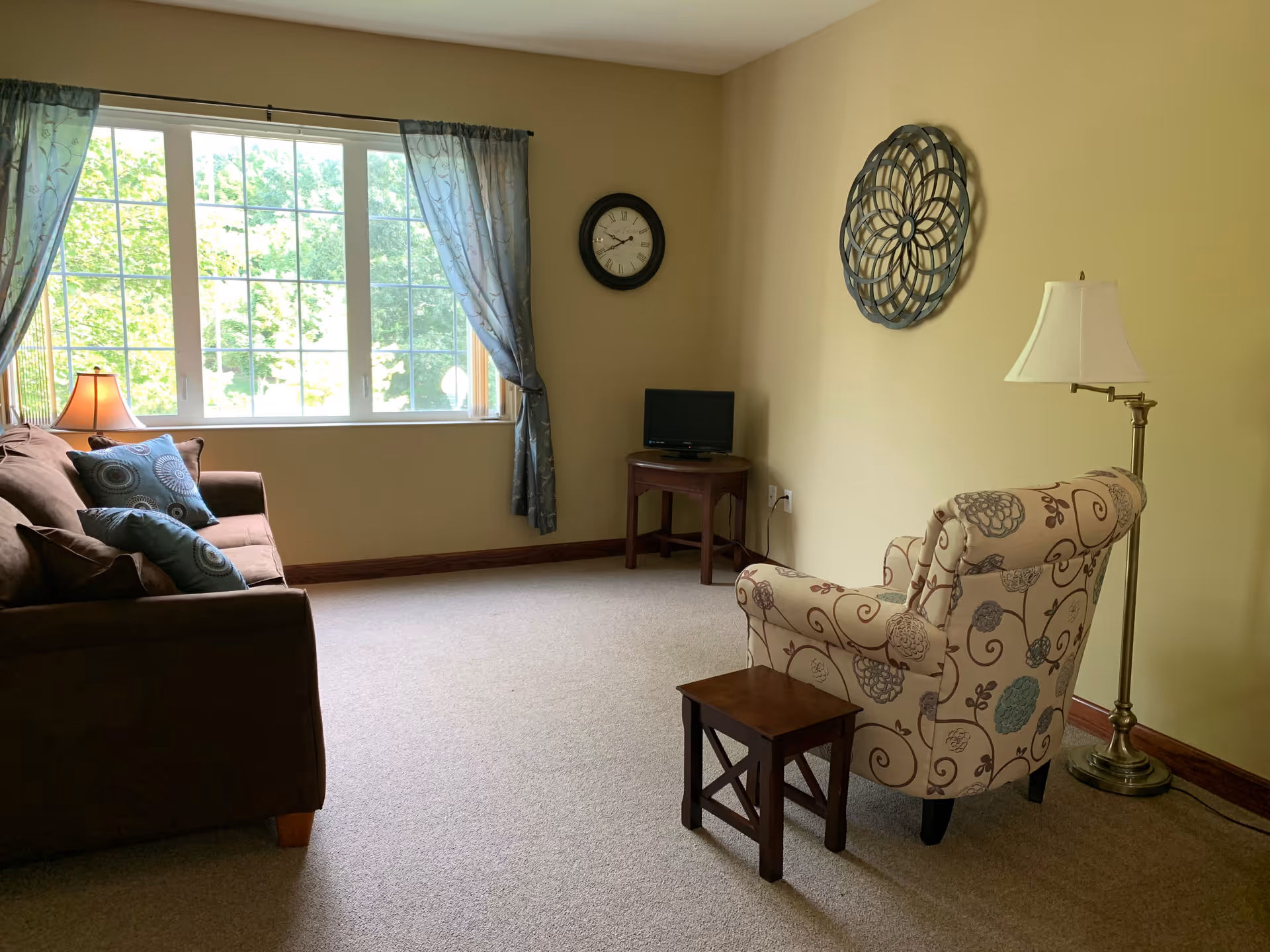 A cozy living room with a sofa by a large window, a patterned armchair, small tables, and a floor lamp.