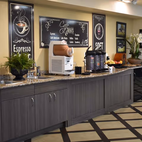 A coffee and refreshment station with a granite countertop and dark wooden cabinets below. On the counter, there is a coffee machine, three large coffee dispensers, a bowl of bananas, and other snacks. The wall behind features decorative signs with coffee-themed artwork and text such as 'Espresso' and 'Coffee Treats'. There are also framed pictures and a potted plant on the counter.