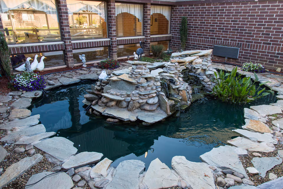 A small outdoor pond with a rock waterfall feature surrounded by flat stones and plants. Decorative statues of ducks and an angel are placed near the pond. The pond is located next to a brick building with large windows.