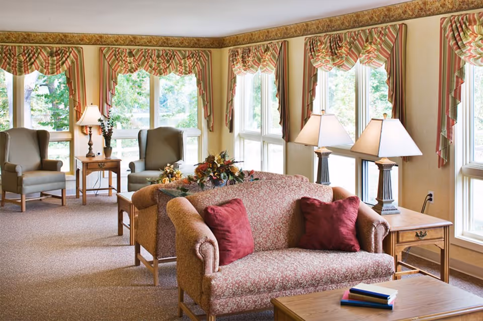 Sunlit communal living room with an upholstered sofa and armchairs arranged by large windows, side tables, lamps, and decorative pillows.
