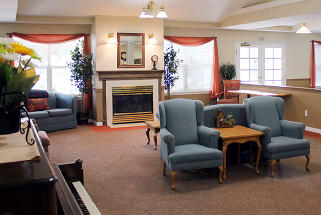 A bright common area with upholstered armchairs and sofas arranged around a fireplace, piano, and side tables.