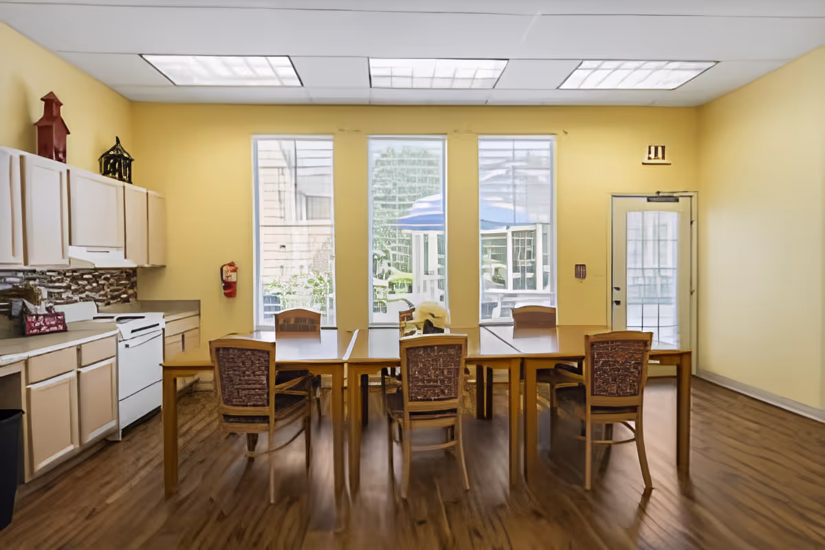 A bright room with yellow walls featuring a kitchen area on the left with white cabinets, a stove, and a countertop. In the center, there are two wooden tables pushed together with six wooden chairs around them. Large windows and a glass door at the back let in natural light and show an outdoor patio with umbrellas and greenery.