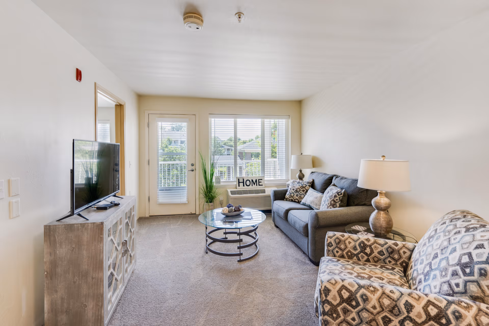 A bright living room with a gray couch adorned with patterned pillows, a matching patterned armchair, a glass coffee table, and a TV on a wooden stand. There is a window with blinds and a door leading to a balcony or outside area. A decorative sign that reads 'HOME' is placed on the windowsill.