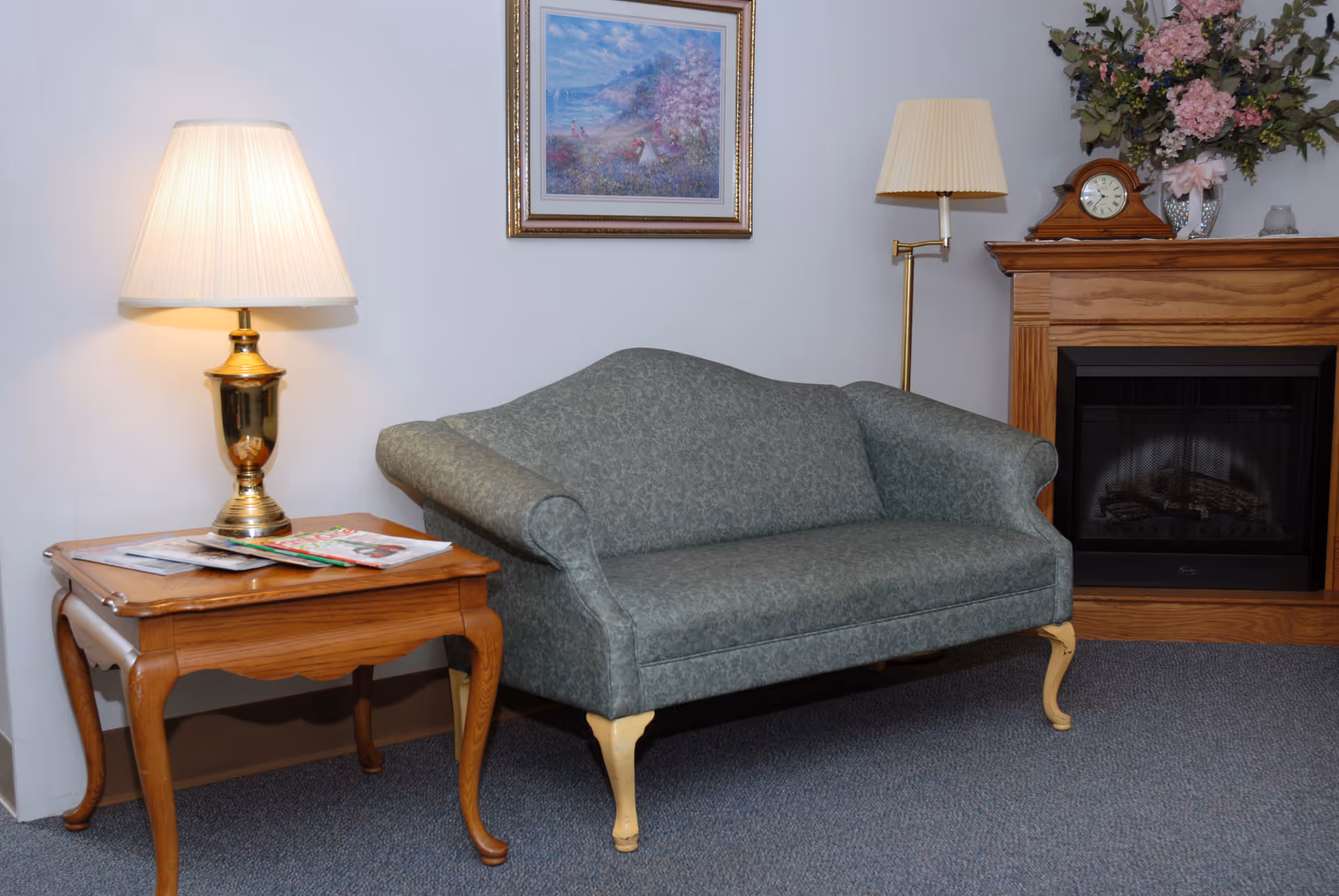Cozy seating area featuring a green upholstered loveseat flanked by a wooden side table with a lamp and a fireplace mantel topped with flowers and a clock.
