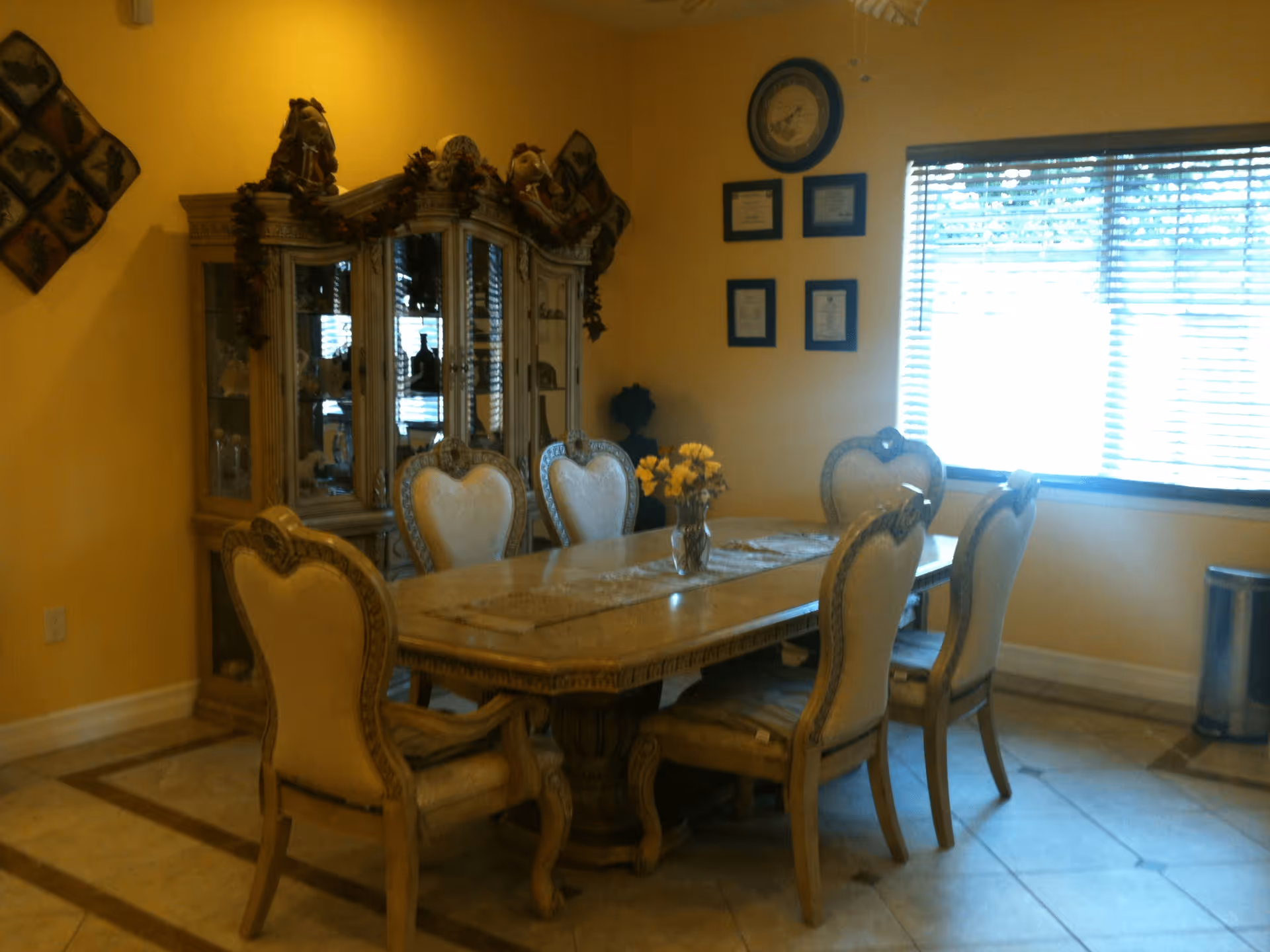 A dining room with a rectangular wooden table surrounded by six ornate chairs with cushioned seats and backs. A vase with yellow flowers is placed on a table runner in the center of the table. Behind the table is a large wooden china cabinet decorated with garlands and various decorative items inside. The walls are painted yellow, and there is a window with blinds letting in natural light. Several framed certificates and a clock are hung on the wall next to the window.