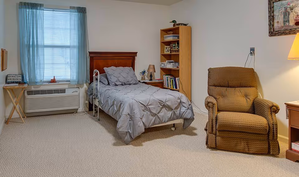 A furnished senior living bedroom with a single bed, recliner, bookshelf, window with blue curtains and an air conditioning unit.