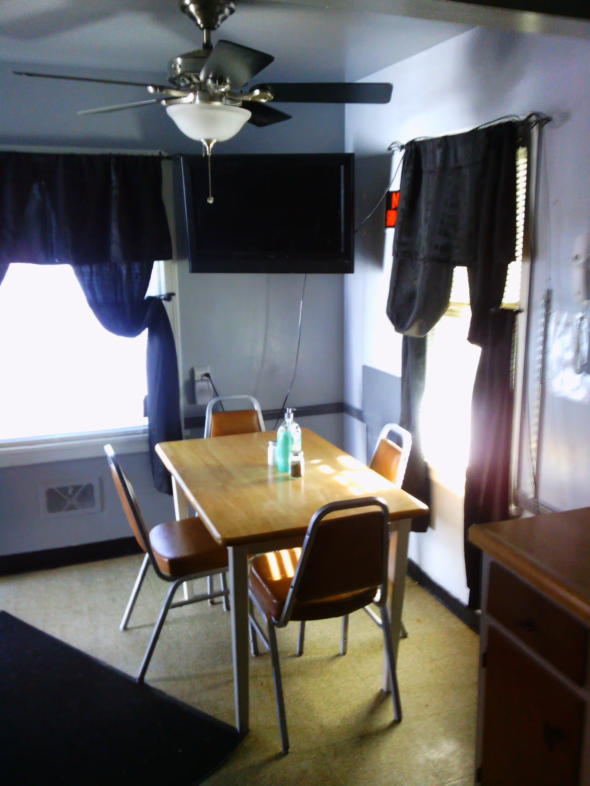 A small dining area with a wooden table and four chairs with brown seats. The room has two windows with dark curtains, a ceiling fan with light, and a wall-mounted flat screen TV. There are some items on the table including a bottle of hand sanitizer and a salt shaker.
