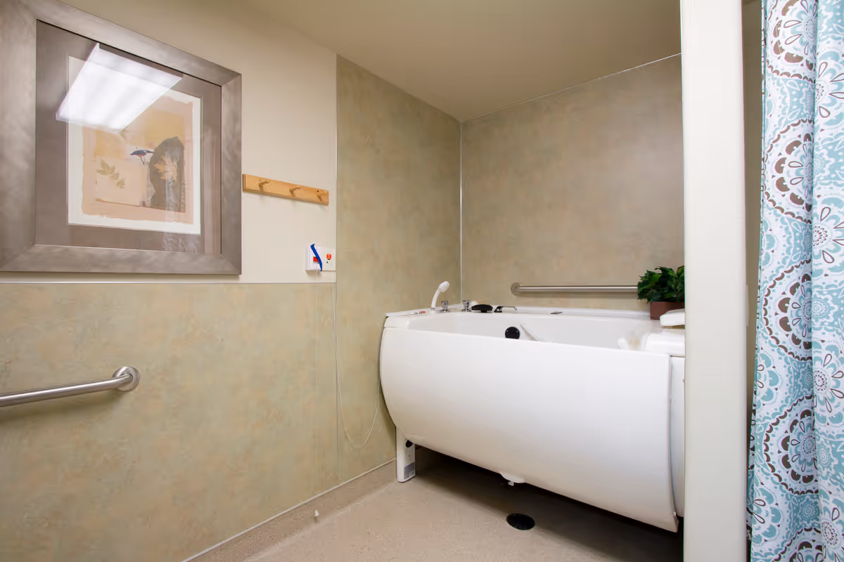 A bathroom with a large white accessible bathtub, grab bars on the walls, a framed picture, a wooden coat rack, and a patterned shower curtain partially visible on the right side.
