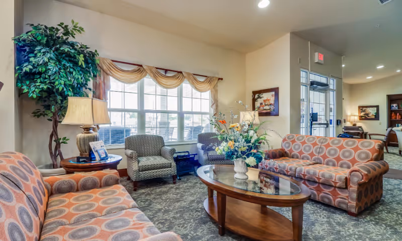 A cozy living room area in an assisted living facility featuring two patterned sofas and two armchairs arranged around a wooden coffee table with a floral centerpiece. A large window with sheer curtains allows natural light to fill the room. There is a tall potted plant and a table lamp on a side table next to one sofa. The room has carpeted flooring and light-colored walls with framed artwork. In the background, there is a glass door entrance and additional seating areas.