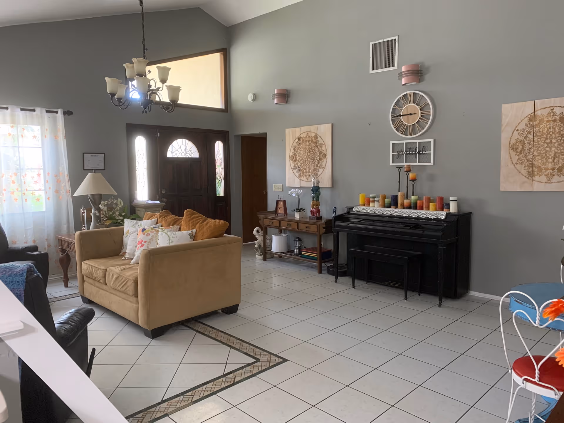 A cozy living room with a beige loveseat adorned with floral pillows, a black piano decorated with colorful candles, a wooden console table with decorative items, a chandelier hanging from a high ceiling, and a large wooden front door with glass panels. The walls are painted gray and decorated with two large circular art pieces and a clock.