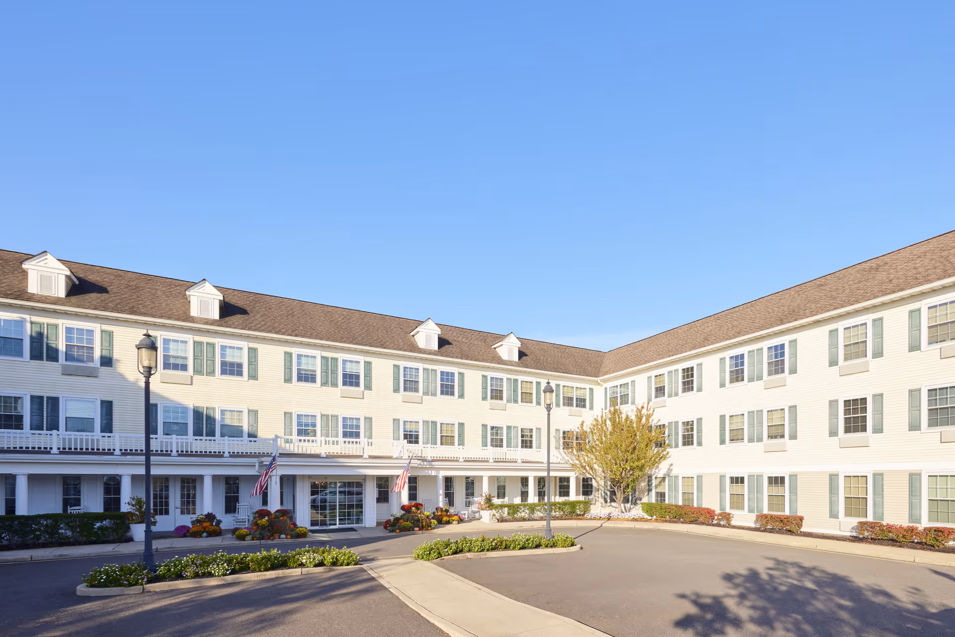 Exterior view of a three-story senior living facility building with white siding, green shutters, and a brown roof under a clear blue sky. The entrance is decorated with flowers and American flags, and there are two street lamps and landscaped greenery in front.