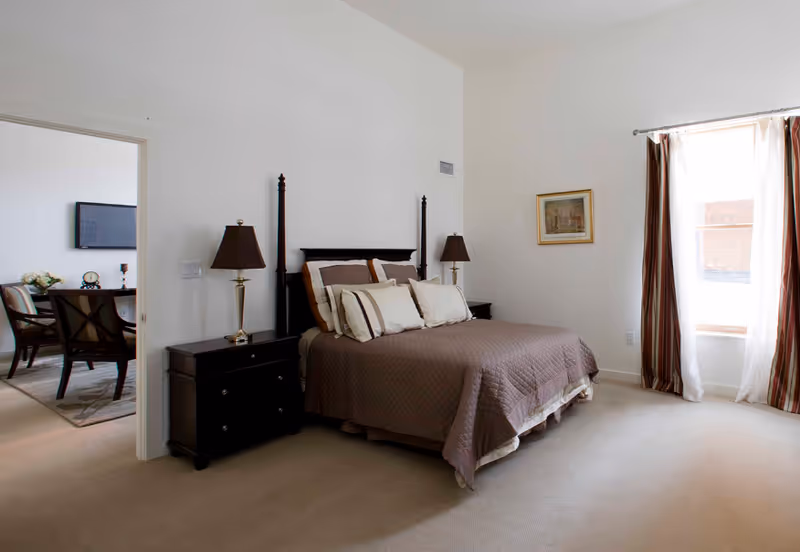 A bedroom with a large bed covered in a brown quilt and multiple pillows. There are two dark wooden nightstands on either side of the bed, each with a lamp. A window with sheer white curtains and brown striped drapes lets in natural light. A framed picture hangs on the wall next to the window. Through an open doorway, a dining area with chairs and a table is visible.
