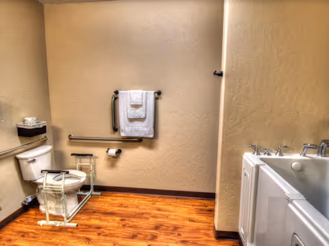 Accessible bathroom with wooden floor, white toilet equipped with safety rails, grab bars on the wall, a towel rack with white towels, and a white bathtub with handrails.