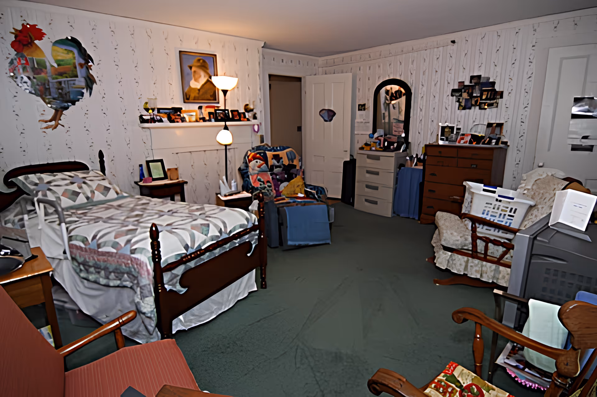 A cozy bedroom in a senior living facility with a single wooden bed covered with a patterned quilt. The room has several chairs, a dresser with a mirror, a chest of drawers, and various personal items and photos displayed. The walls are decorated with wallpaper and a painting, and there is a floor lamp providing light.