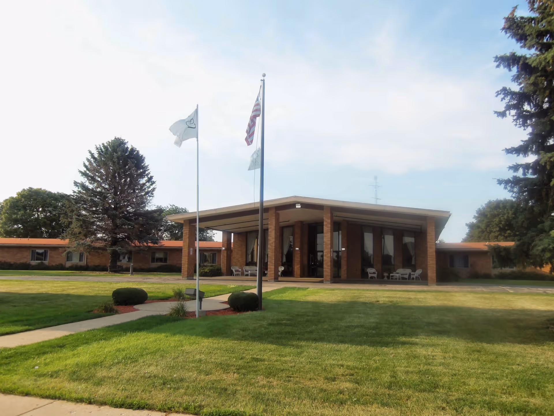 Front entrance of a single-story brick senior living facility with flagpoles and a manicured lawn.
