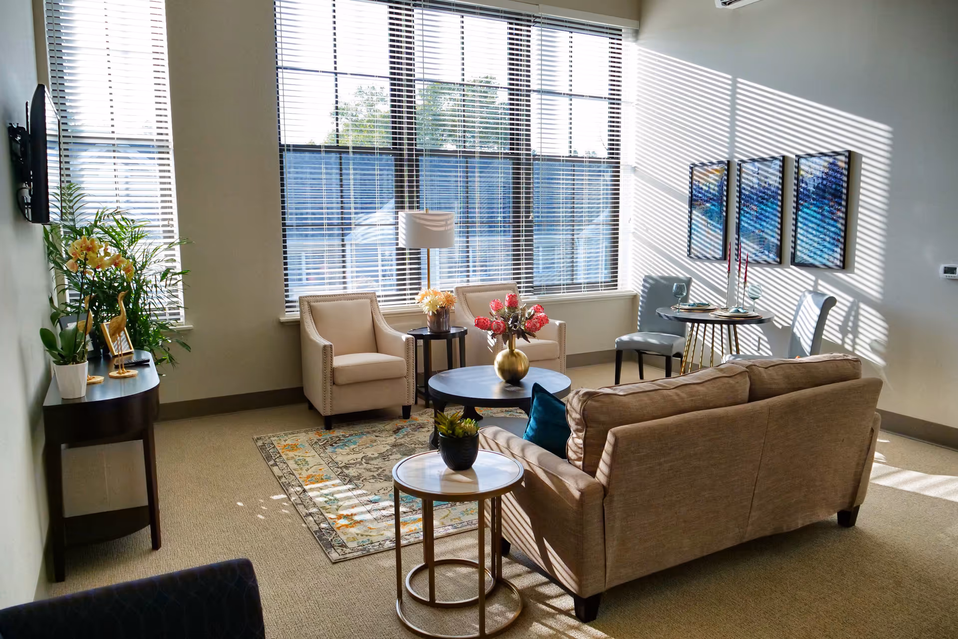 Sunlit common living room with a sofa, armchairs, coffee tables, plants and wall art in front of large windows.