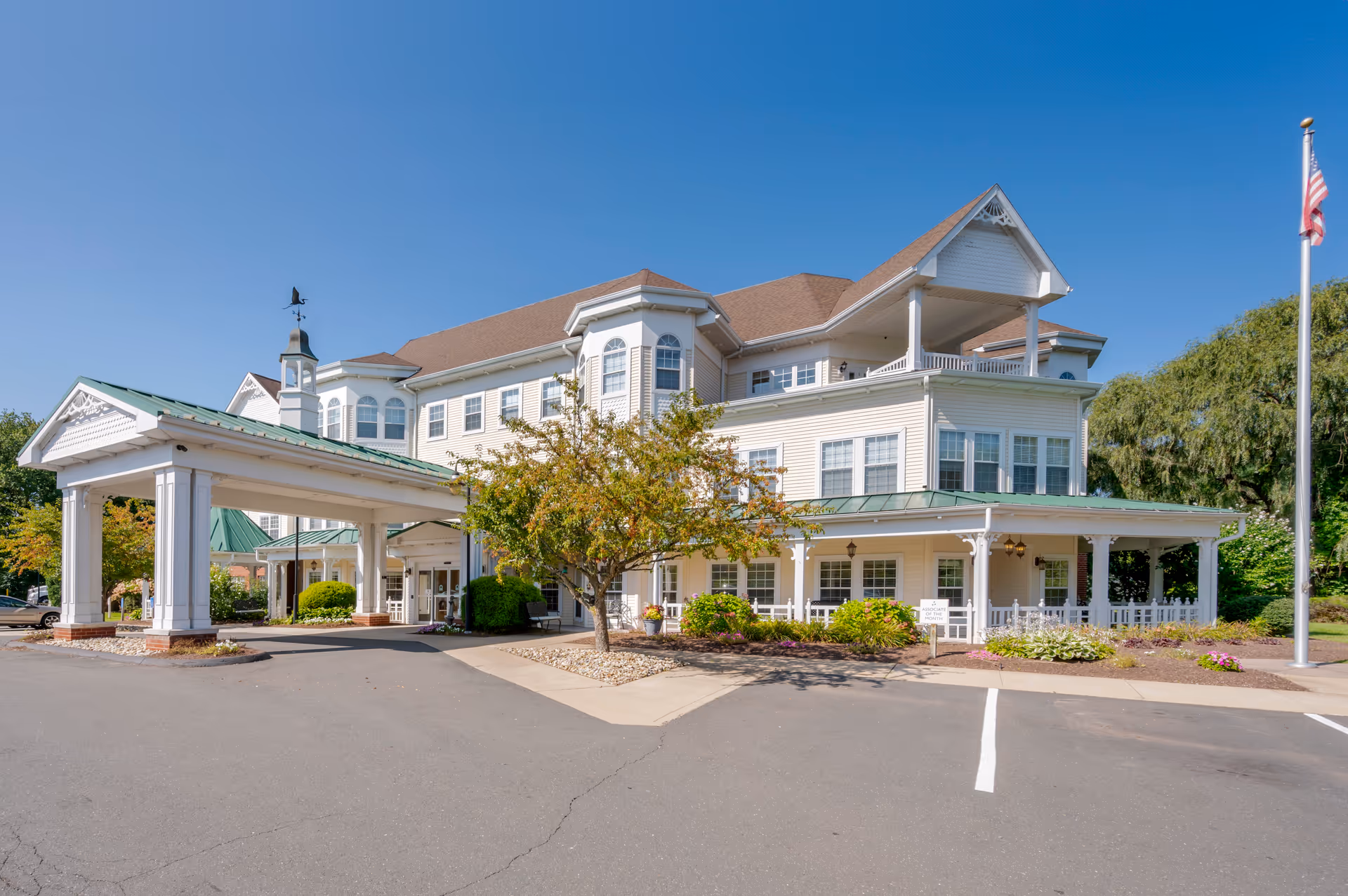 Exterior view of a large, multi-story senior living facility with white siding and a brown roof. The building features a covered entrance with white columns, a small tree and landscaped bushes in front, and an American flag on a flagpole to the right. The sky is clear and blue.