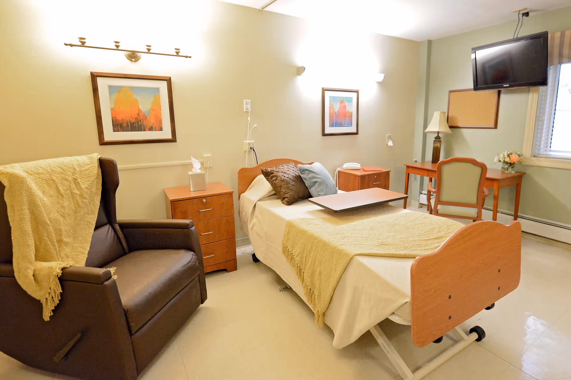 A senior living bedroom with an adjustable bed, recliner, dresser, desk, and wall-mounted TV.