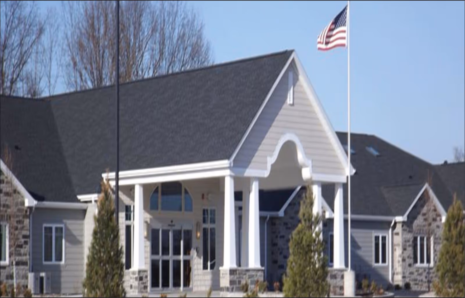 Front entrance of a single-story assisted living building with a covered portico and an American flag.