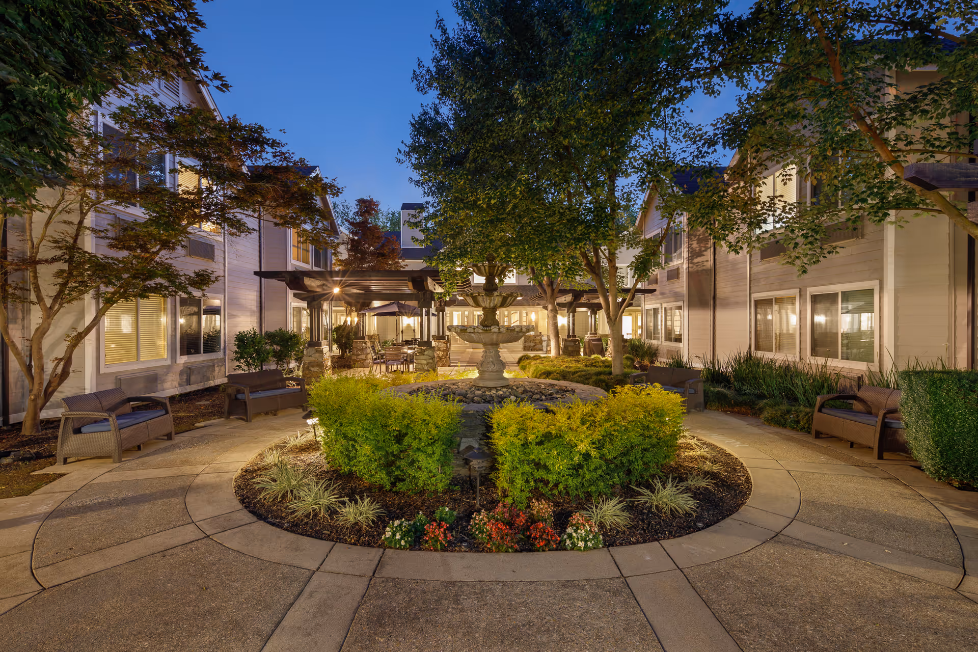 Outdoor courtyard area at dusk with a central multi-tiered stone fountain surrounded by green bushes and flowers. There are curved concrete walkways with benches on either side, trees providing shade, and a pergola with seating and tables in the background. The courtyard is flanked by two-story residential buildings with lit windows.