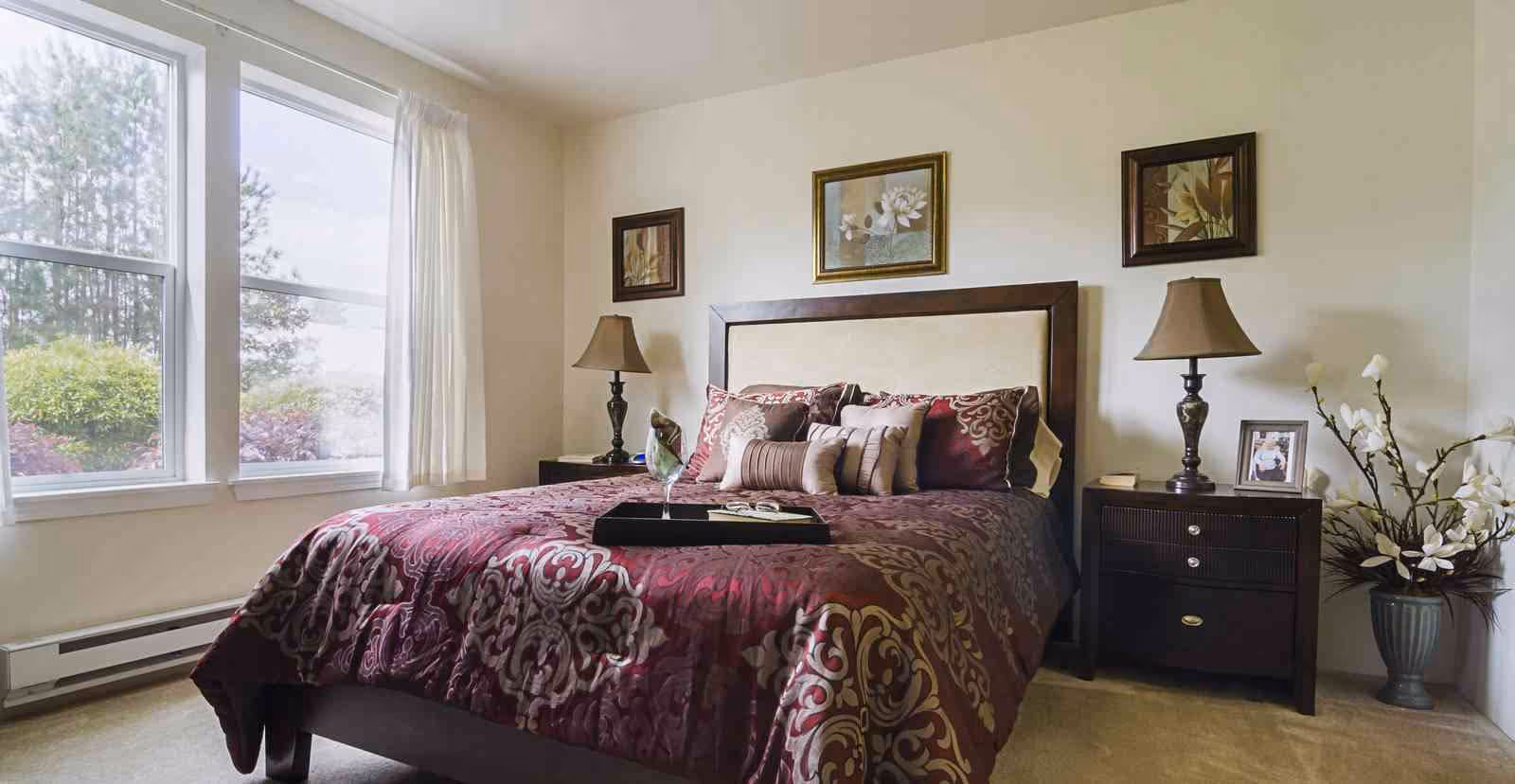 Sunlit bedroom with a queen bed dressed in patterned burgundy bedding, two nightstands with lamps, framed artwork above the headboard, and a window showing greenery outside.
