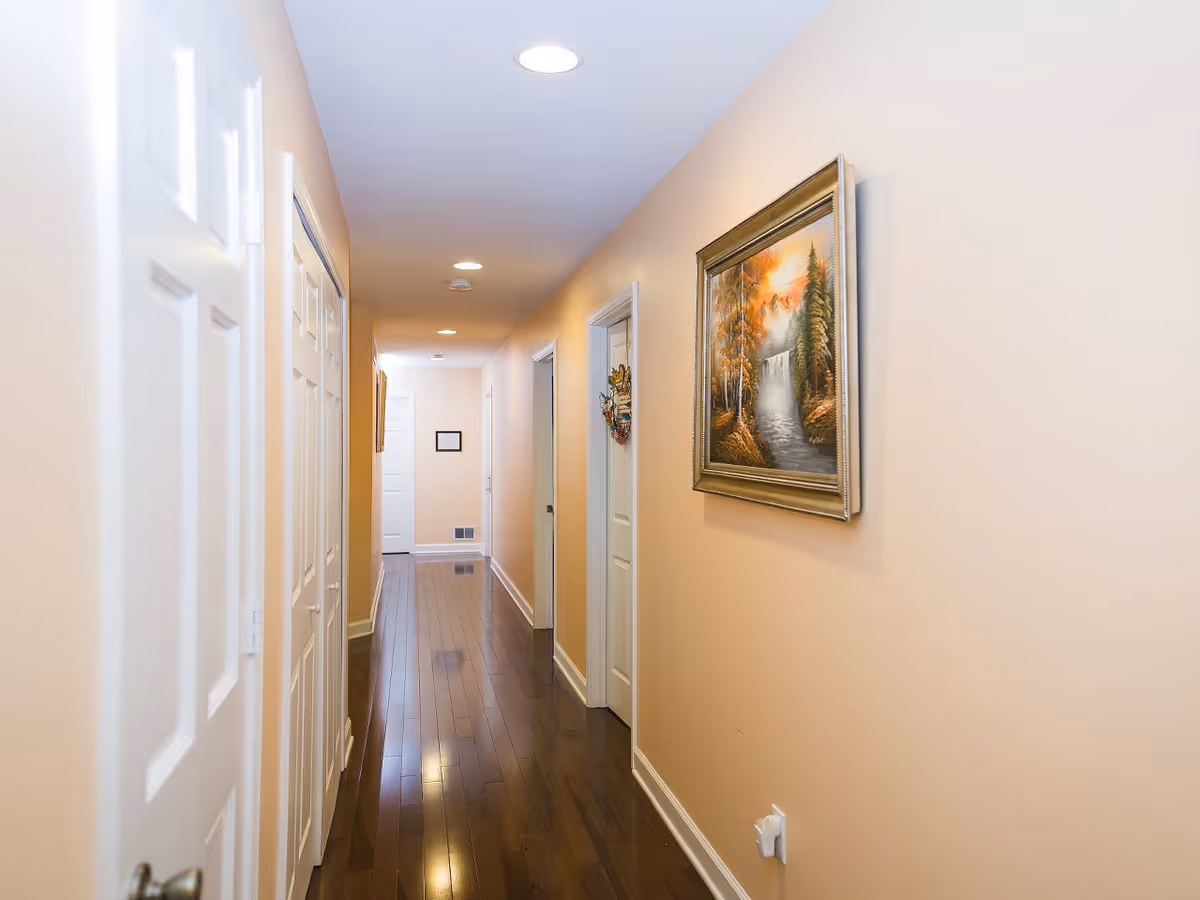 A long, narrow hallway with light beige walls and dark wooden flooring. Several white doors line both sides of the hallway, and a framed painting of a waterfall and trees hangs on the right wall. Recessed ceiling lights illuminate the space.