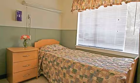 A small bedroom in a senior facility with a single bed, dresser, and window.