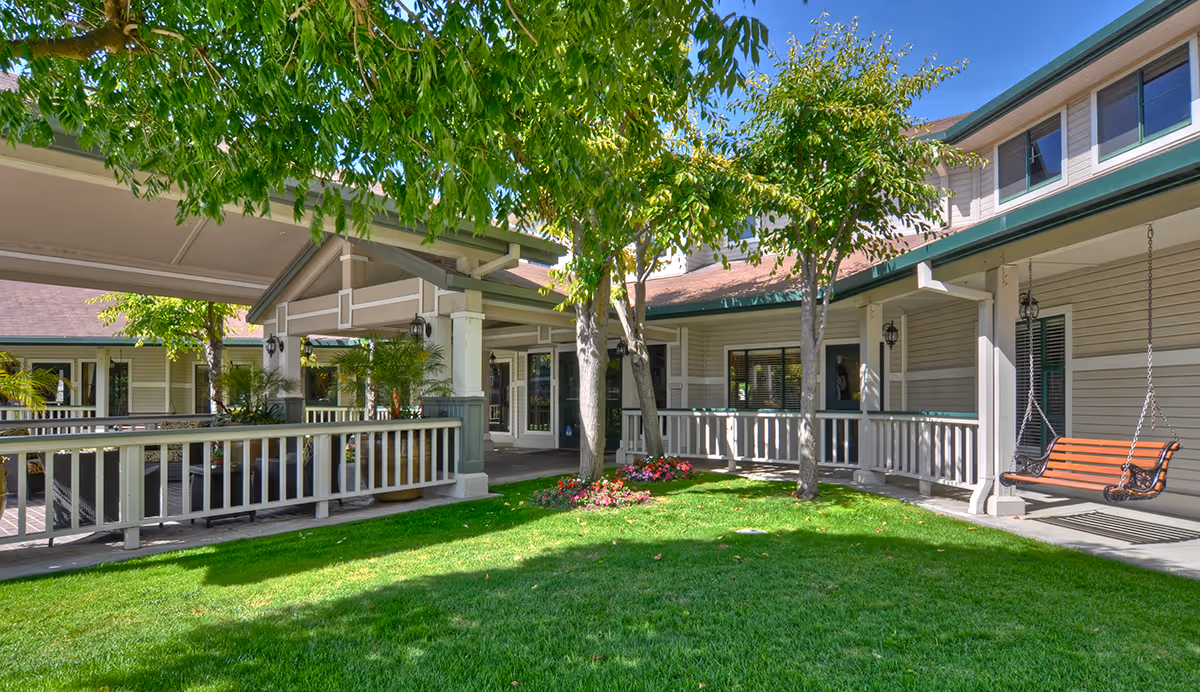 Outdoor courtyard area at Ivy Park at Salinas featuring green grass, trees, a flower bed, a covered walkway with white railings, and a wooden swing bench hanging from the ceiling.