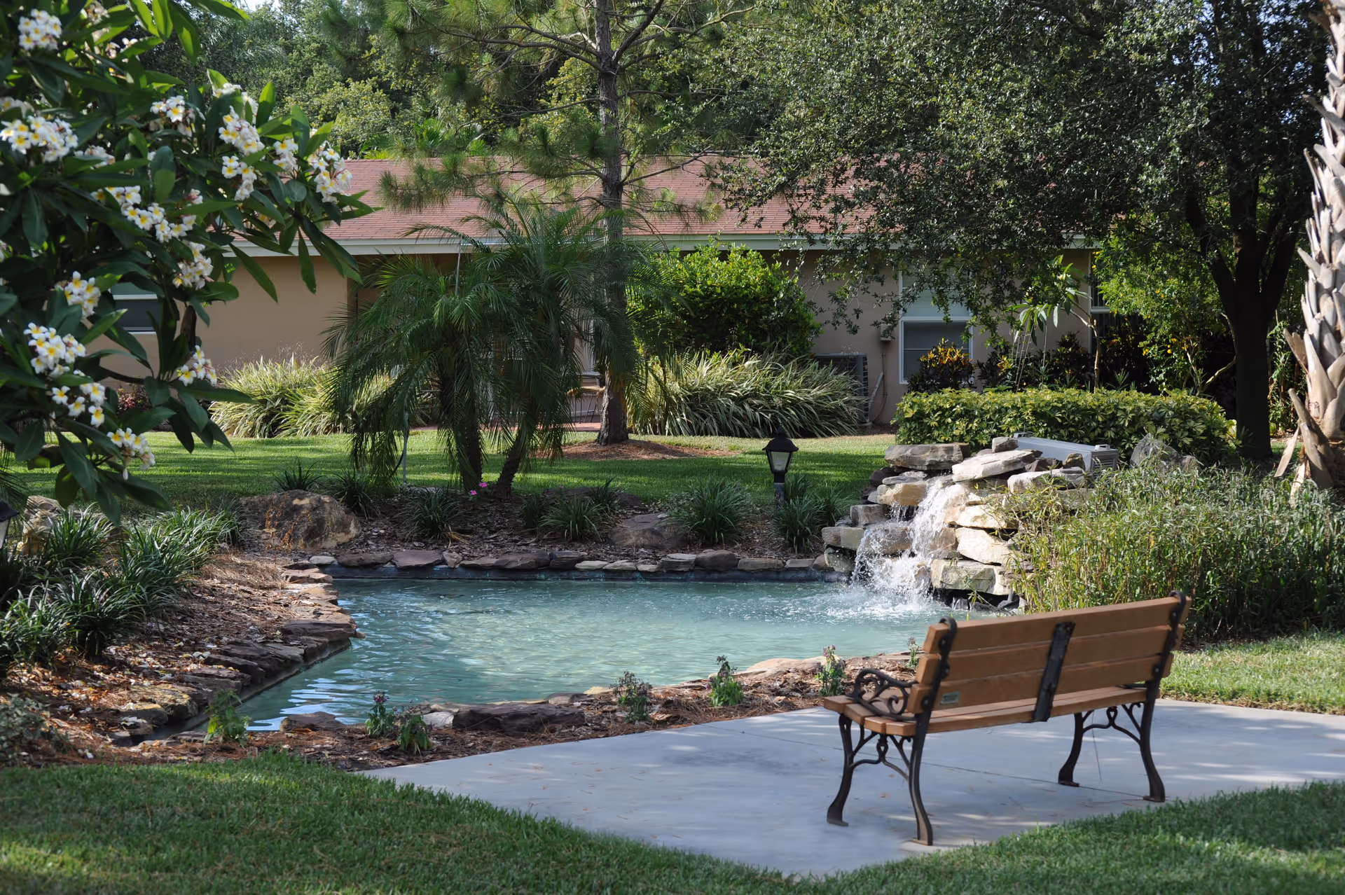 A landscaped outdoor area with a small pond and rock waterfall, a bench on a paved path, and greenery with a building in the background.
