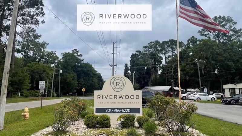 Outdoor view of the entrance sign for Riverwood Health and Rehab Center in Starke, FL, with a landscaped area featuring bushes and rocks in front of the sign. An American flag is flying on a flagpole to the right, and there are trees, power lines, and parked cars in the background under a cloudy sky.