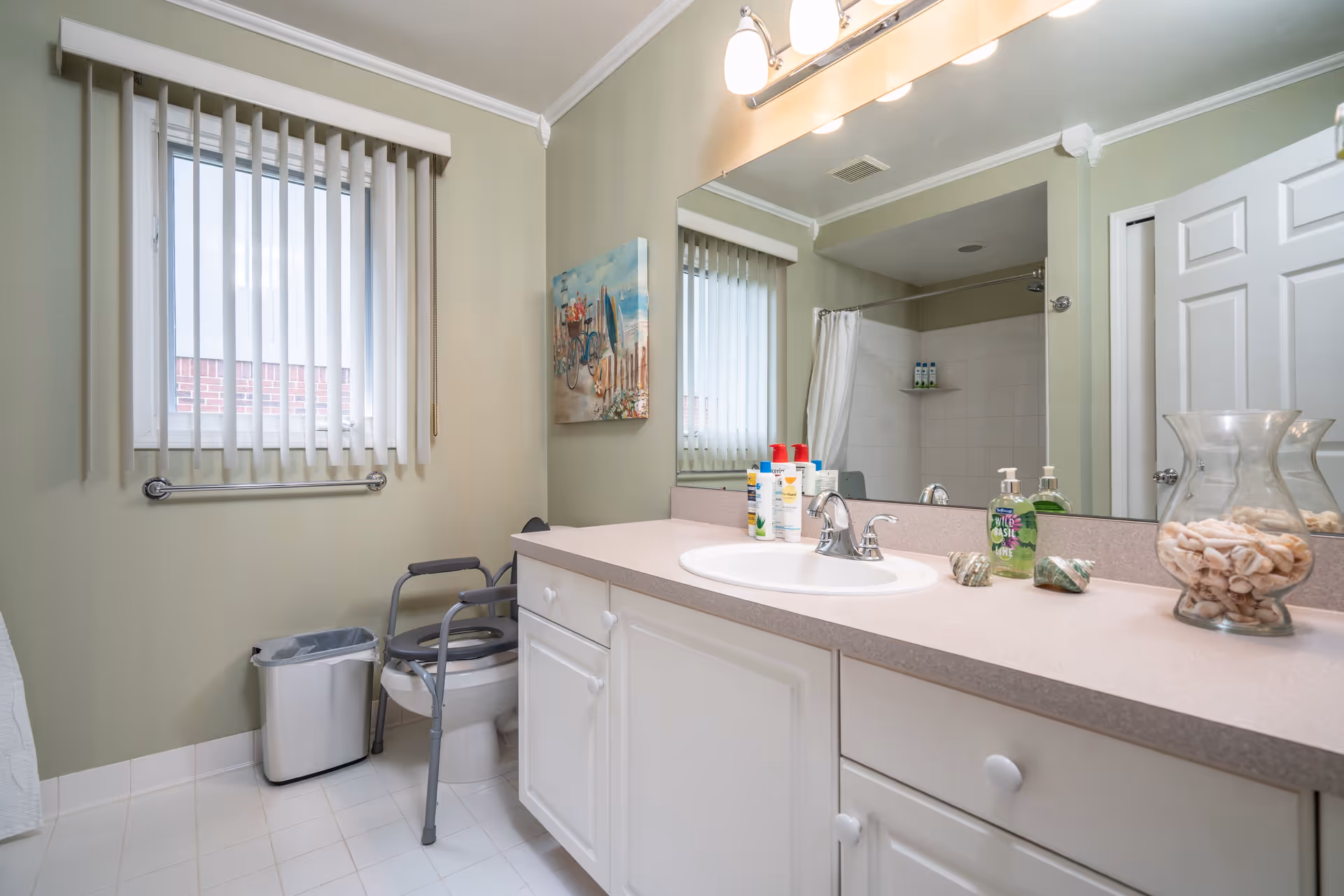 A bathroom with a white vanity and sink, a large mirror above the sink, and a window with vertical blinds. There is a toilet with a gray safety frame around it, a trash can, and a shower with a white curtain. Various toiletries and decorative seashells in a glass vase are on the countertop. The walls are painted light green.
