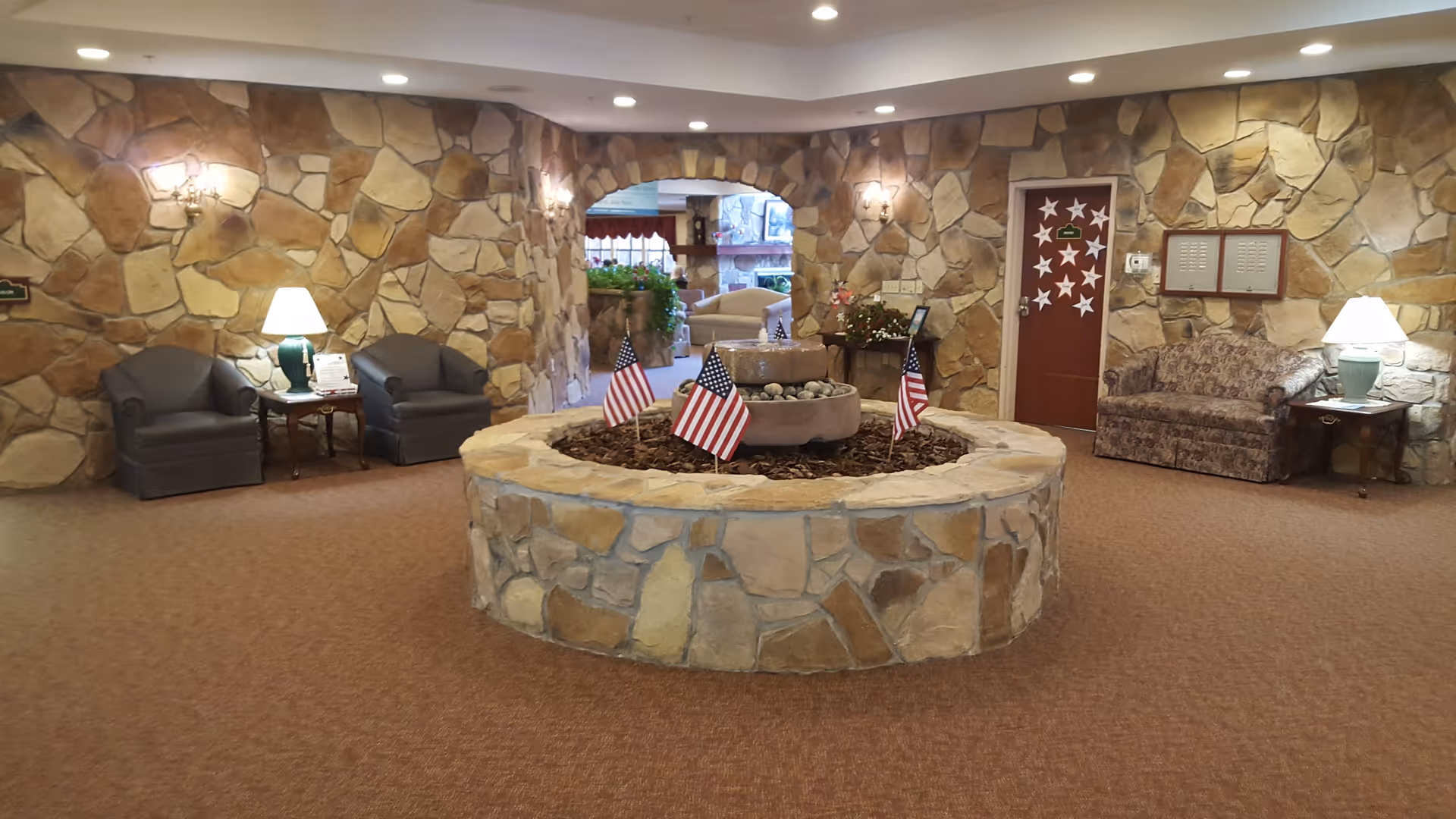 Interior of a senior living facility with stone walls and carpeted floor. In the center, there is a circular stone planter with a small water fountain and several American flags. Surrounding the planter are seating areas with armchairs and lamps on side tables. A doorway decorated with star cutouts is visible on the right side of the image.