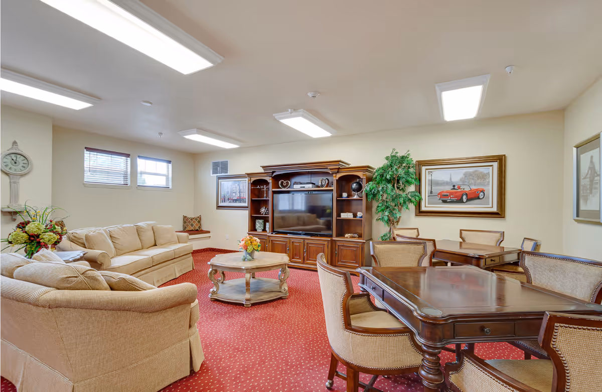 A cozy living room area in a senior living facility featuring beige upholstered sofas, a wooden coffee table with a flower arrangement, and a large wooden entertainment center with a flat-screen TV. The room has red carpet with a subtle pattern, several framed pictures on the walls, a large potted plant, and a wooden table with chairs in the foreground. The space is well-lit with ceiling lights and has two small windows with blinds.