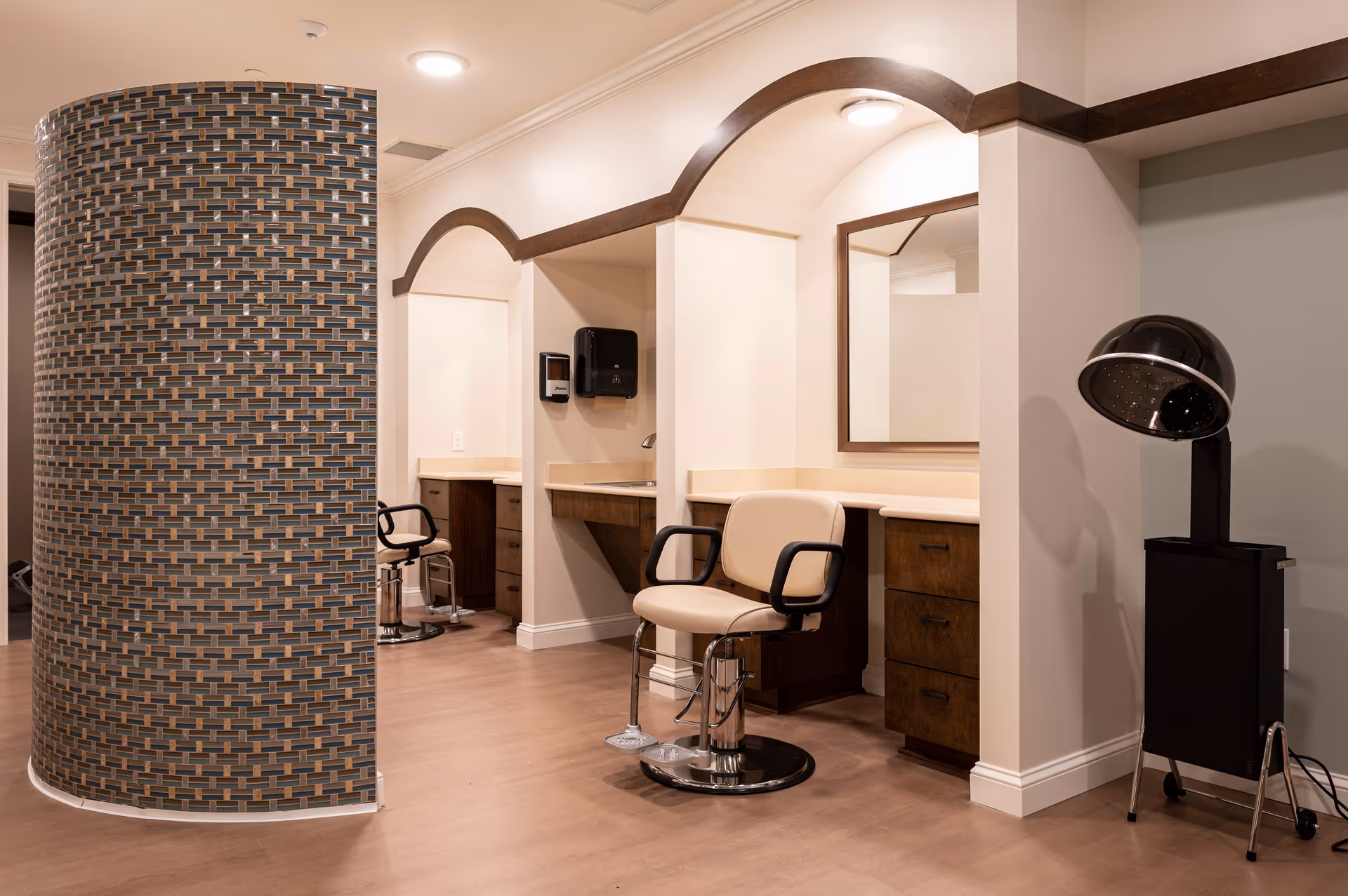 Interior view of a senior living facility salon area with two styling stations featuring chairs, mirrors, and wooden cabinetry. A curved wall with mosaic tiles partially separates the space, and a black hair dryer hood on a stand is visible on the right side.